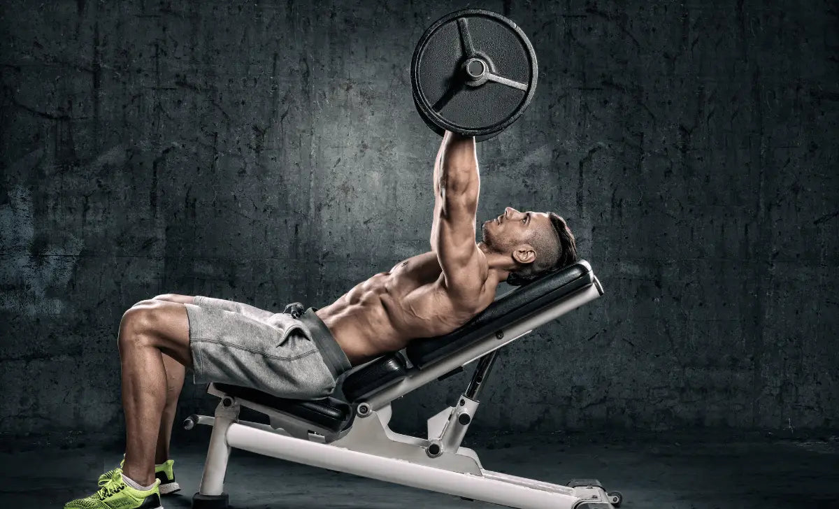 Homme musclé effectuant un développé incliné à la barre sur un banc de musculation, sur fond de mur en béton.